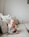 A baby sits on a beige couch holding and chewing on a mushie Linen Pacifier Clip 2-Pack in a bright, minimalist living room.