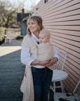 A woman smiles outdoors by a pink wall, holding a baby in a beige wrap. The alert baby uses a FRIGG Natural Rubber pacifier from the FRIGG 6-Pack while sitting near white chairs and a table on this sunny day.