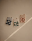 Three mushie Phone Press Toys in beige, gray, and blush, each with six circular buttons, are displayed on a light brown surface with diagonal sunlight—ideal for sensory play and fine motor skill development.
