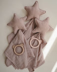 Three mushie Star Lovey Blankets in soft mauve rest on a light surface beside two silicone teething rings, shaped like a bear and cat. Sunlight casts gentle shadows to the right.