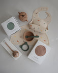 A flat lay of baby feeding essentials featuring a beige animal-print bib, a mushie Silicone Placemat, food-grade silicone utensils, a snack cup, teether, and two boxed silicone bibs in green and pink on a light background.