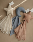 Three mushie Star Lovey Blankets made from organic cotton rest on a beige surface: one with a cream star, one with a blue moon, and one with a dusty pink star. Each features matching fabric and a tag at the top.