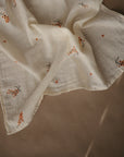The mushie Organic Cotton Muslin Cloths 3-Pack features light, cream-colored fabric with orange floral prints and a subtle plaid texture—soft, delicate, and ideal for nurseries or diaper bags. Draped on a brown surface.
