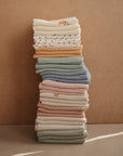 The mushie Organic Cotton Muslin Cloths 3-Pack features cozy essentials in cream, mustard, sage, blue, and blush—perfect for nursery needs. Two neat stacks rest against a neutral beige background.