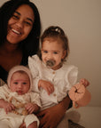 A smiling woman holds a baby in white with a knit bonnet and a toddler in a white shirt using the FRIGG Moon Silicone Pacifier 2-Pack by FRIGG, surrounded by baby essentials in a softly lit room.