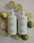Two pale green pump-top bottles of mushie Green Lemon Baby Body Lotion, made with natural ingredients and featuring a fresh scent, are displayed among whole and halved green lemons on a light background.