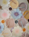 A party tabletop features large paper flowers and pastel mushie Round Dinnerware Plates, cups, and forks in soft pink, yellow, lavender, and blue. The non-toxic plates ensure safety while adding playful charm to the decor.