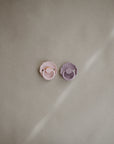 Two FRIGG Daisy Natural Rubber Pacifiers—one pink and one purple—are placed side by side on a beige surface with a diagonal light streak in the background.