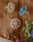 Four pastel FRIGG Andersen Fairytale Natural Rubber Pacifiers (2-packs) are displayed on wood, with green leaves and purple flowers in the corner and a white cloth to the left.