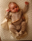 A baby in a beige outfit lies on the mushie Extra Soft Muslin Changing Pad Cover with neutral polka dots, wearing a beaded pacifier clip. Warm, gentle lighting adds to the cozy scene.