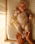 A baby in a beige outfit and knit hat lies on the mushie Extra Soft Muslin Changing Pad Cover, holding a pacifier. An adult's hands gently touch the baby, with a tube of lotion nearby as sunlight streams in.