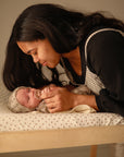 A woman smiles warmly while gently offering a pacifier to a baby in a knitted hat, lying on the mushie Extra Soft Muslin Changing Pad Cover.