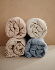 Four rolled extra soft mushie muslin changing pad covers are stacked in two rows on a beige background. The covers come in soft tones: beige, cream, blue, and beige with white stripes.