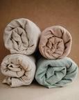Four rolled muslin blankets in cream, beige, light blue, and stripes are stacked together against a plain brown background, resembling the mushie Extra Soft Muslin Changing Pad Cover.