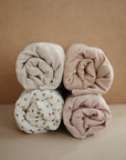 Four rolled-up swaddles in neutral tones sit in a square—three solid beige and brown, one white with brown florals—beautifully matching your mushie Extra Soft Muslin Changing Pad Cover against a beige background.