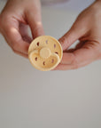 A person holds a FRIGG Moon Silicone Pacifier from the FRIGG brand above a white surface, showcasing this beige, medical-grade silicone baby essential from the 2-Pack set.