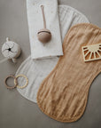 On a gray surface: mushie's Organic Cotton Muslin Burp Cloth 2-Pack (beige, white), a dotted muslin cloth, silicone snack cup, pacifier clip, two teething rings, and a sun-shaped toy in neutral tones.