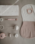 A flat lay of soft neutral baby items, including mushie's Organic Cotton Muslin Burp Cloth 2-Pack, a bib, pacifier clip, teether, rattles, silicone toy, and pacifier—all neatly arranged on a beige surface.