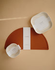 A set of minimalist, BPA-free square dinnerware plates by mushie in beige and rust, along with a bowl and semicircular placemat, is displayed on a neutral tan background.