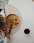 A child with glasses sits at a table, eating a heart-shaped waffle served on a mushie Round Dinnerware Plate. Three raspberries and a fork are on the plate, with a cup nearby on the white surface. The view is from above.