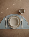 The mushie Round Dinnerware Plates, plus a beige spoon, fork, bowl, and cup are arranged on a blue half-circle placemat atop a brown tabletop; all pieces are minimalistic and dishwasher safe.