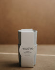 Two stacked light gray "mushie" Dinnerware Cups, each with a white label reading “mushie CUPS dishwasher safe made in Denmark.” Made of non-toxic plastic, these kids cups rest on a neutral light brown surface.