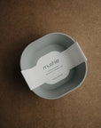 A light gray, square mushie Square Dinnerware Bowl with rounded edges and a BPA-free design sits on a brown surface. A white label with "mushie DINNER BOWL" in minimalist font is across the bowl.