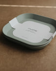 A light green mushie Square Dinnerware Plate with rounded edges and BPA-free material rests on a brown surface, featuring the brand's name at the center and a white paper band with text wrapped around it.