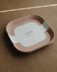 The mushie Square Dinnerware Plates set includes two BPA-free pink plastic plates with rounded edges, stacked together and wrapped in a white label marked mushie, displayed on a brown textured surface.