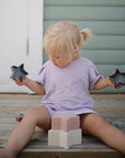 A young child with blonde pigtails, in a lavender shirt, sits on wooden steps holding the mushie Nesting Stars Toy and looks down at stacked blocks. A light blue wall is in the background.