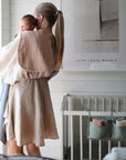 A woman with a ponytail holds a baby over her shoulder in a softly lit nursery, standing beside a crib with storage pockets and mushie Organic Cotton Muslin Burp Cloth 2-Pack. A modern art print decorates the wall.