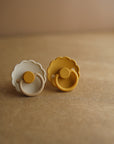 The FRIGG Daisy Natural Rubber Pacifier 2-Pack by FRIGG includes one beige and one mustard yellow scalloped-edge pacifier, each with a gentle natural rubber latex nipple, shown side by side on a brown textured surface.