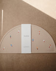 A beige, semi-circular Silicone Placemat by mushie, made from food-grade silicone and featuring colorful rocket illustrations. It comes with a white mushie-branded band and is shown on a brown surface with a diagonal shadow.