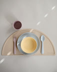 A pastel kids place setting features mushie Round Dinnerware Plates with a pink fork, yellow bowl, blue spoon, and mauve cup on a beige semicircular placemat—all dishwasher safe and neatly arranged.