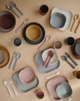 Mushie Round Dinnerware Plates in earthy, muted tones are neatly arranged on a beige surface, forming a symmetric pattern with their non-toxic plastic design.