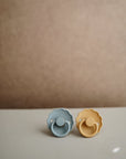 The FRIGG Daisy Natural Rubber Pacifier 2-Pack by FRIGG features daisy-shaped blue and yellow pacifiers with scalloped edges and natural rubber latex nipples, displayed side by side on a softly lit surface.