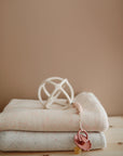 Two folded beige baby blankets rest on a wooden surface, topped with a white geometric rattle and mushie's Silicone Pacifier Clip | Hera. A pacifier is attached to the clip. The plain background is a soft brown wall.