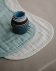 A stack of colorful nesting cups rests on mushie's Organic Cotton Muslin Burp Cloth 2-Pack in soft blue and cream, displayed on a neutral beige surface.