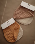 Two sets of mushie baby bibs sit on a brown surface, each pairing a solid brown or mustard bib with a cream shade—ideal to coordinate with the mushie Organic Cotton Muslin Burp Cloth 2-Pack.