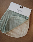 The mushie Organic Cotton Muslin Burp Cloth 2-Pack, featuring one sage green and one cream cloth, is shown on a brown surface.