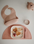 A mushie Square Dinnerware Plate, sippy cup, and fork in pink are displayed with heart-shaped pancakes, banana slices, and raspberries on a pink speckled placemat against a white background.
