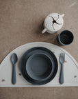 A minimalist mushie Round Dinnerware Bowl set in gray and beige, BPA-free, includes baby bowls, plate, spoon, fork, cup, snack cup, and a semicircular placemat with rainbow designs on a brown surface.