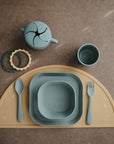 The mushie Square Dinnerware Plates set includes BPA-free silicone and plastic baby plates, cup, bowl, utensils, snack cup, and teething rings in muted blue and mustard—stylishly arranged on a brown surface.