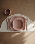 The mushie Square Dinnerware Plates set, BPA-free and in pink, including a plate, bowl, cup, spoon, and fork, is neatly placed on a cream half-moon placemat atop a brown surface and viewed from above in natural light.