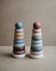 Two mushie Stacking Rings Toys, pastel-colored and non-toxic, form cone shapes topped with round balls and are placed side by side on a neutral textured background.
