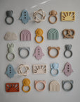 A neatly arranged grid of mushie Sun Teether and other colorful, non-toxic silicone baby teethers in fun shapes—rockets, rainbows, clouds, animals, pretzels—displayed on a neutral background.