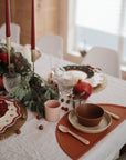 A festive dining table features red candles, greenery, pomegranates, decorative plates, and mushie Round Dinnerware Plates on a tan placemat. The non-toxic, dishwasher safe plates add a practical touch to the bright scene.