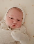 A newborn rests calmly on the mushie Extra Soft Muslin Changing Pad Cover in cream with small sun designs, dressed in a light hooded outfit and mittens.