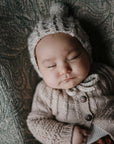 A baby peacefully sleeps, wearing a cream-colored knitted hat and cardigan, lying on a mushie Extra Soft Muslin Changing Pad Cover featuring a green patterned design.