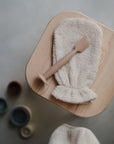A brown Mushie Diaper Cream Applicator and a beige exfoliating mitt sit on a wooden stool, with small muted round containers placed on the floor nearby.